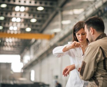 Beautiful female medic bandaging injured worker's hand at industrial factory hall.